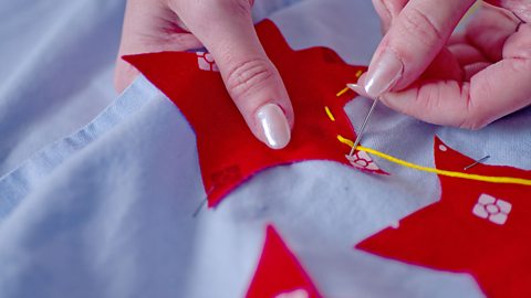 A close up of a woman's hands holding a fabric garment while threading yellow thread through to attach a red fabric star