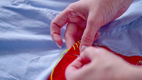 A woman's hands threading yellow thread around the top of a red fabric pocket with a needle