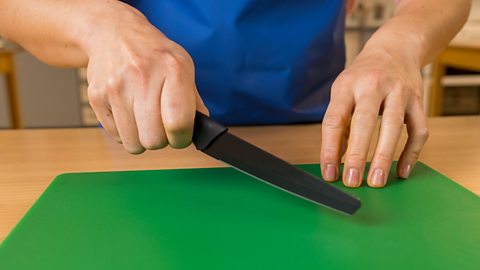 A woman's hand holding a black knife downwards, with the other hand resting on a green chopping board