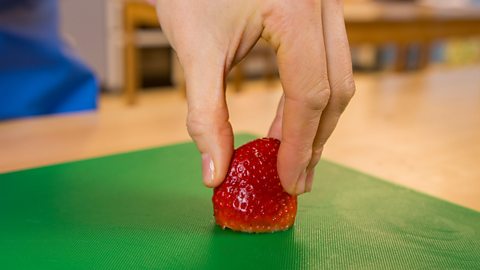 A woman's hand holding a strawberry on a green chopping board, between her thumb and fingers