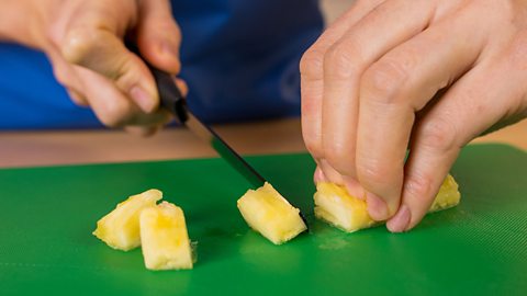 A woman's hands with a knife and several small pieces of cut pineapple on a green chopping board