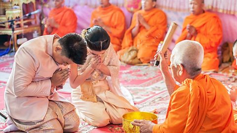 A photo of three people; a couple on the left with heads bowed in prayer face a Buddhist monk on the right during a wedding blessing.