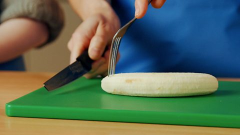 A child holding a fork above a banana and a knife in their writing hand.