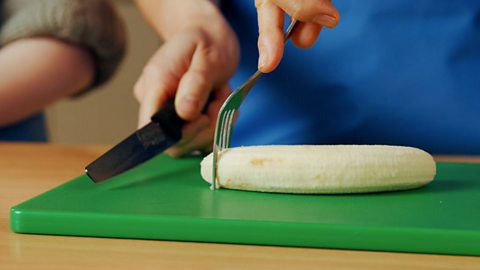 The fork cutting pressed into the banana and the child ready to make a slice