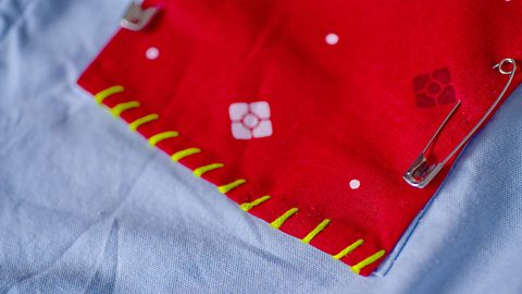 A close up of a red fabric pocket attached to a blue shirt, with yellow stitches threaded over the top edge