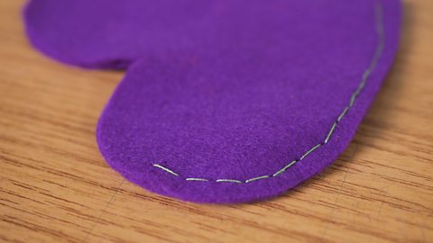 A purple felt heart laying on a wooden table, with a line of closely sewn stitches around the edge