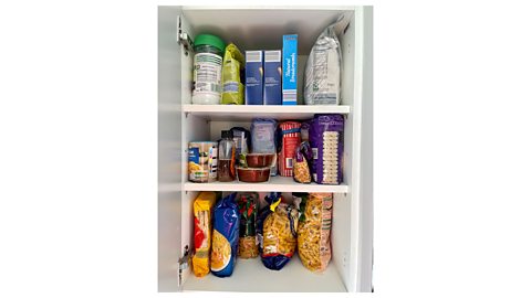 A kitchen cupboard with three shelves, containing a selection of dried foods