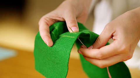 A pair of hands holding a piece of green felt and pushing a needle up through the back of the fabric