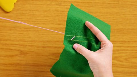 A pair of hands holding a piece of green felt and pulling pink thread through to make a diagonal stitch