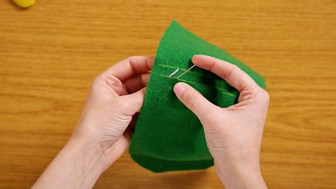 A pair of hands holding a piece of green felt and pushing a needle up through the fabric in line with an existing stitch