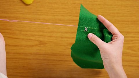 A pair of hands holding a piece of green felt and pushing a needle up through the fabric , crossing over the first stitch, making a cross