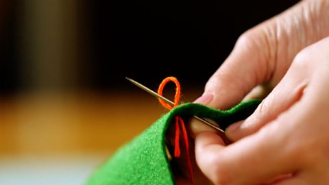 A pair of hands holding green fabric and pushing a needle up through a loop of red thread