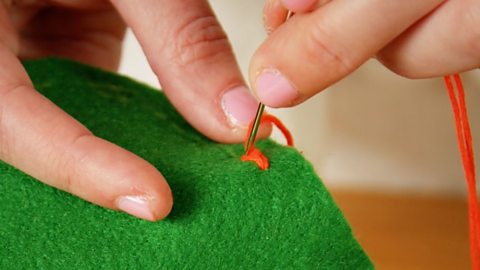 A pair of hands holding green fabric and pushing a needle down through a closed loop of red thread