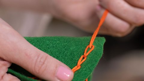 A pair of hands holding green fabric and pushing a needle through the end loop of a line of stitches