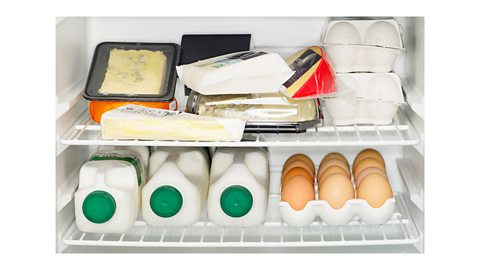 An inside view of a fridge with packets of cheese and eggs on the top shelf, and three plastic bottles of milk with green tops and eggs on the lower shelf