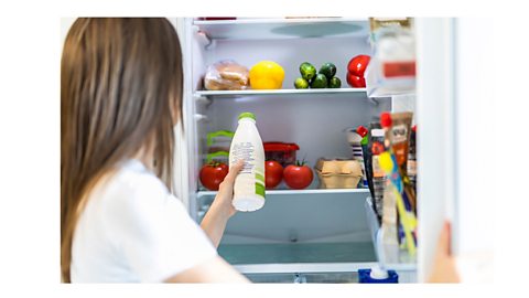 A woman taking out a bottle of milk from a fridge, with tomatoes, lemons, eggs and other soft focus fridge items