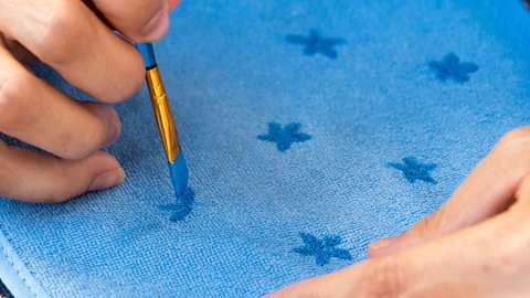 A pair of hands using a paintbrush to paint blue flowers onto blue fabric