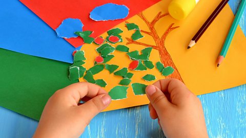 A child's hands holding small green paper pieces and putting them onto yellow card with a tree drawn on. Two colouring pencils and a glue stick is on the the table