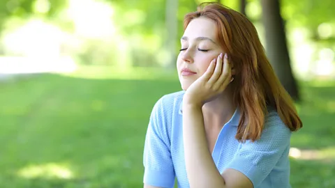 A woman sits with her eyes closed in a sunny, green space (Credit: Alamy)