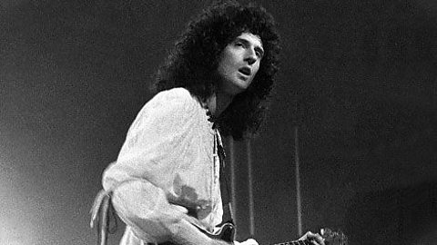 A black and white photo of a male musician wearing a loose white top playing an electric guitar on a stage.