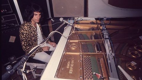 A male wearing white and a gold casual jacket sat at a piano with the lid off showing the strings and hammers inside. 