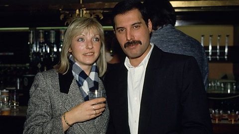 A female and male posed together, smiling, in front of a bar.  The female is holding a drink in her hand,