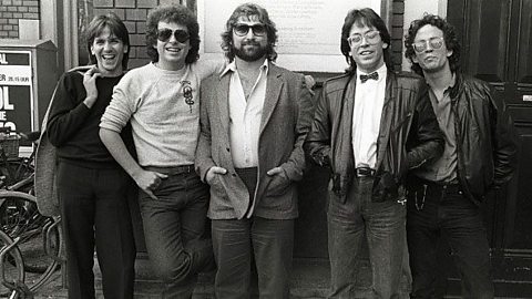 Five men, in smart casual dress posing in a group against a brick wall.   