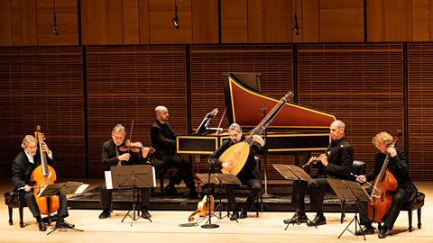 Six musicians sat  behind music stands on a stage all dressed in black suits.  From left to right they are playing a viol, a violin, a harpsichord, a theorbo, a flute and a bass viol.