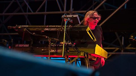 A man wearing sunglasses sat behind a synthesiser performing on a stage.