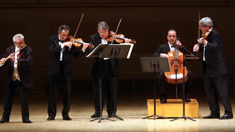 5 musicians performing on stage dressed in black suits.  4 are standing and one is seated all behind music stands.  From left to right there is a flautist, 2 violinists, a cellist and another violinist.