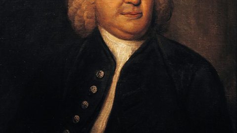 An oil portrait painting of Johann Sebastian Bach. He is wearing a white curly wig and a ruffled white shirt with a jacket with buttons down the left side.  He is holding a piece of paper with musical score on it.  