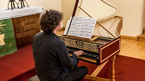 A man playing a harpsichord in a church. Sheet music can be seen on the stand of the instrument and the top is propped open so the strings are in view.