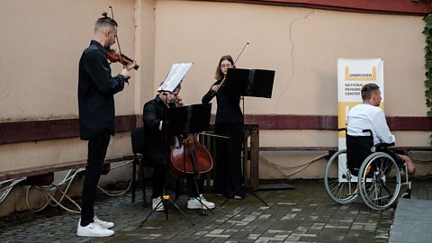 Three musicians busking on a street.  Two standing, one playing a violin and the other a viola.  The third is seated in the middle playing a cello.