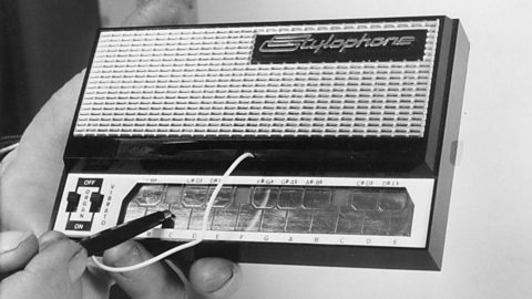 A black and white image of a person holding a small rectangular device with the word Stylophone in the top right corner.  Along the bottom of the box is a metal plate which the person is touching a stylus to.