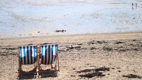 A sandy beach with two blue and white striped deckchairs facing the sea.  The shadow of two people can be seen sitting in the deckchairs and in the top right corner two people are in view playing in the sea.