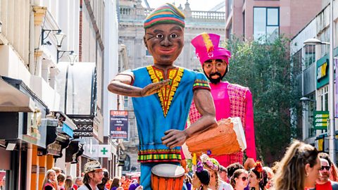 Two processional giants playing drums whilst walking alongside participants in Belfast's multicultural Mela Carnival.