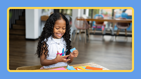 A child using a pair of scissors to cut various coloured bits of paper.