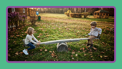Two children playing on a wooden see saw in a park.