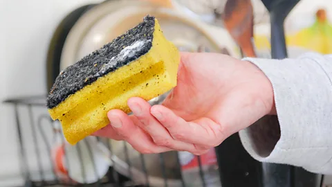 A yellow sponge with a green scourer on top flecked with dirt being held by a person's hand in front of some dishes drying in a rack in the background (Credit: Getty Images)