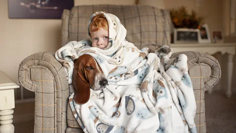 Boy and dog cuddled up together under a blanket on a sofa (Credit: Getty)