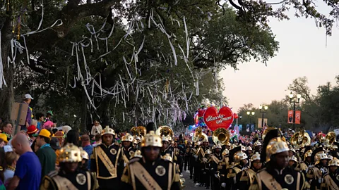 A marching band performs on the streets of New Orleans, with a crowd spectating and streamers in the trees above (Credit Abdul Aziz)