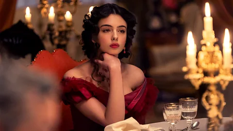 Still from Netflix's The Leopard, showing a close-up girl in red dress sat at a table, with a candelabra in the foreground (Credit: Lucia Iuorio/ Netflix)