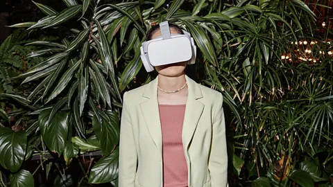 Woman in pink shirt and white blazer wearing VR goggles in front of leafy plants (Credit: Getty Images)