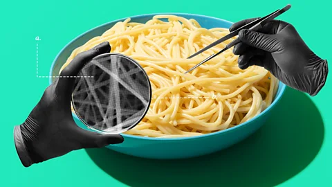 A bowl of pasta with two gloved hands inspecting the noodles with tweezers and a circular scope (Credit: Serenity Strull/ Getty Images)