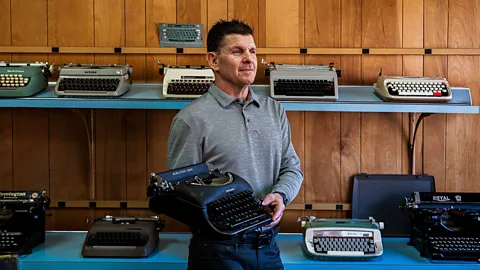 A man in a grey polo-shirt – Mike Marr – proudly holds a typewriter in his hands, with several other models of typewriters on the shelf behind him (Credit: Ernesto Roman)