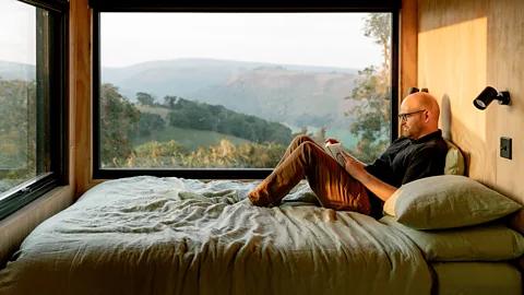 A man sits on a bed reading a book in a cabin which has views out to green hills (Credit: Adam Firman)