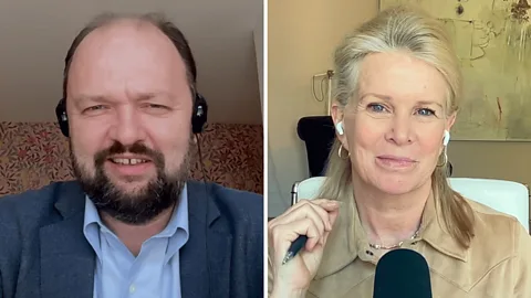 BBC Special Correspondent Katty Kay speaks to author Ross Douthat on a video call. Kay is shown from behind with white earbuds and a black microphone and she wears a tan shirt. Douthat is on her monitor and can be seen wearing a dark blue jacket and black headphones (Credit: BBC)