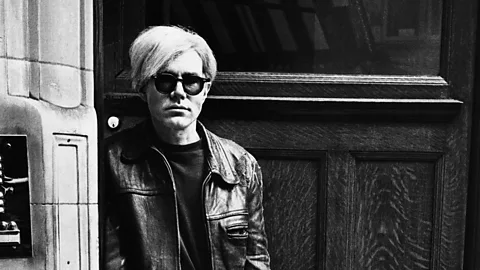 A black and white photo of Andy Warhol wearing sunglasses and leaning against a wall (Credit: Getty Images)