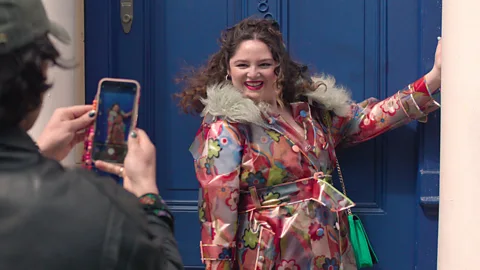 A still of Megan Stalter posing for a photo in front of a blue door in Too Much (Credit: Netflix)