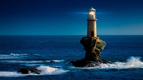 Tourlitis Lighthouse stands on top of a rocky outcrop on the Aegean Sea. Waves crash around it as the light shines on top.